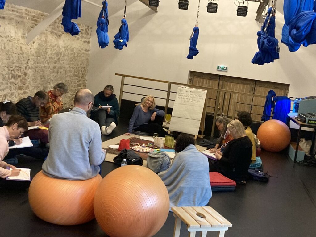 Plusieurs personnes dans une salle avec des hamacs suspendus, des ballons de gym et des tapis. Ils discutent ensemble en cercle